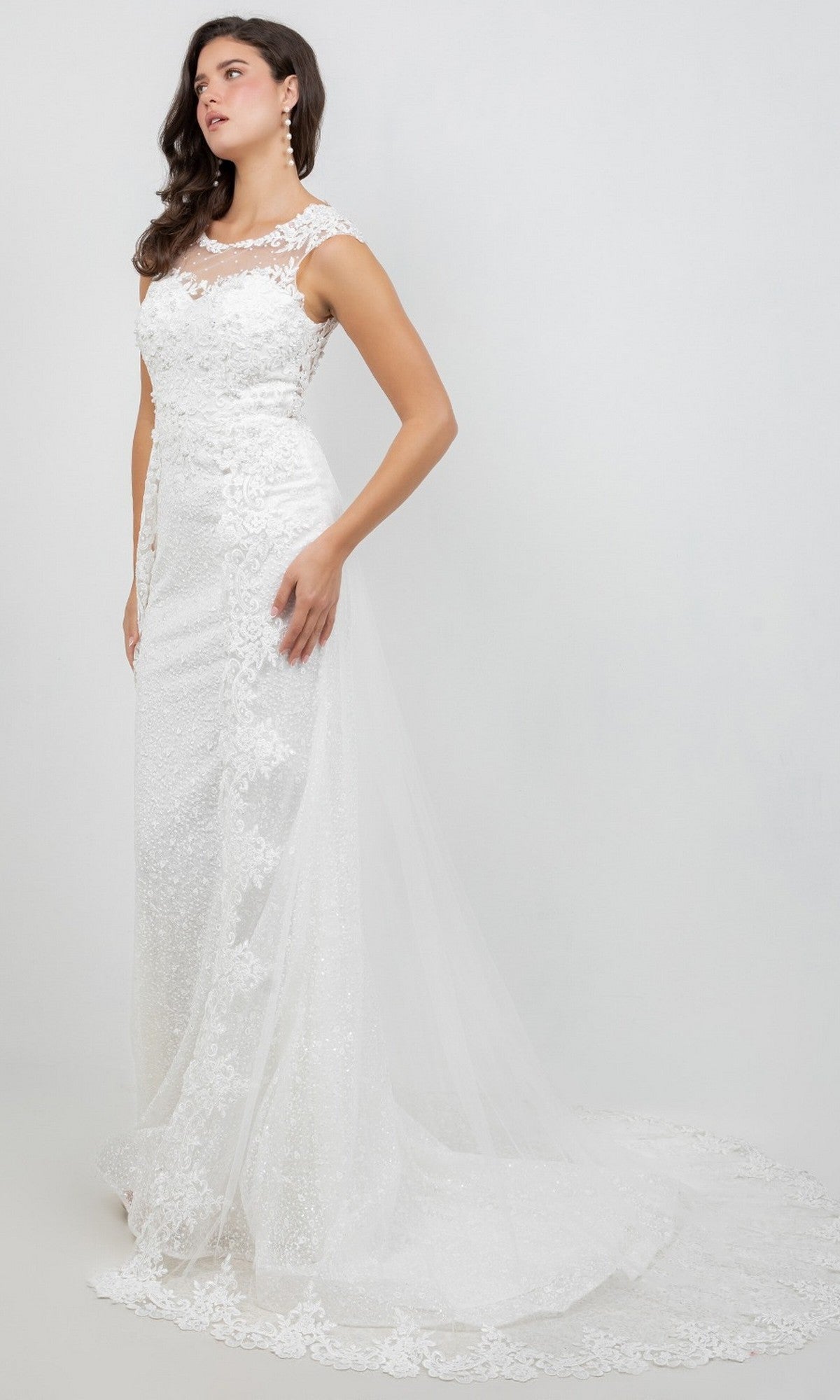 A woman wearing the Cinderella Couture 8506J long formal dress, featuring a sleeveless white lace design with a fitted bodice, illusion neckline, and a sweeping tulle train adorned with intricate lace appliques, stands against a plain light background.